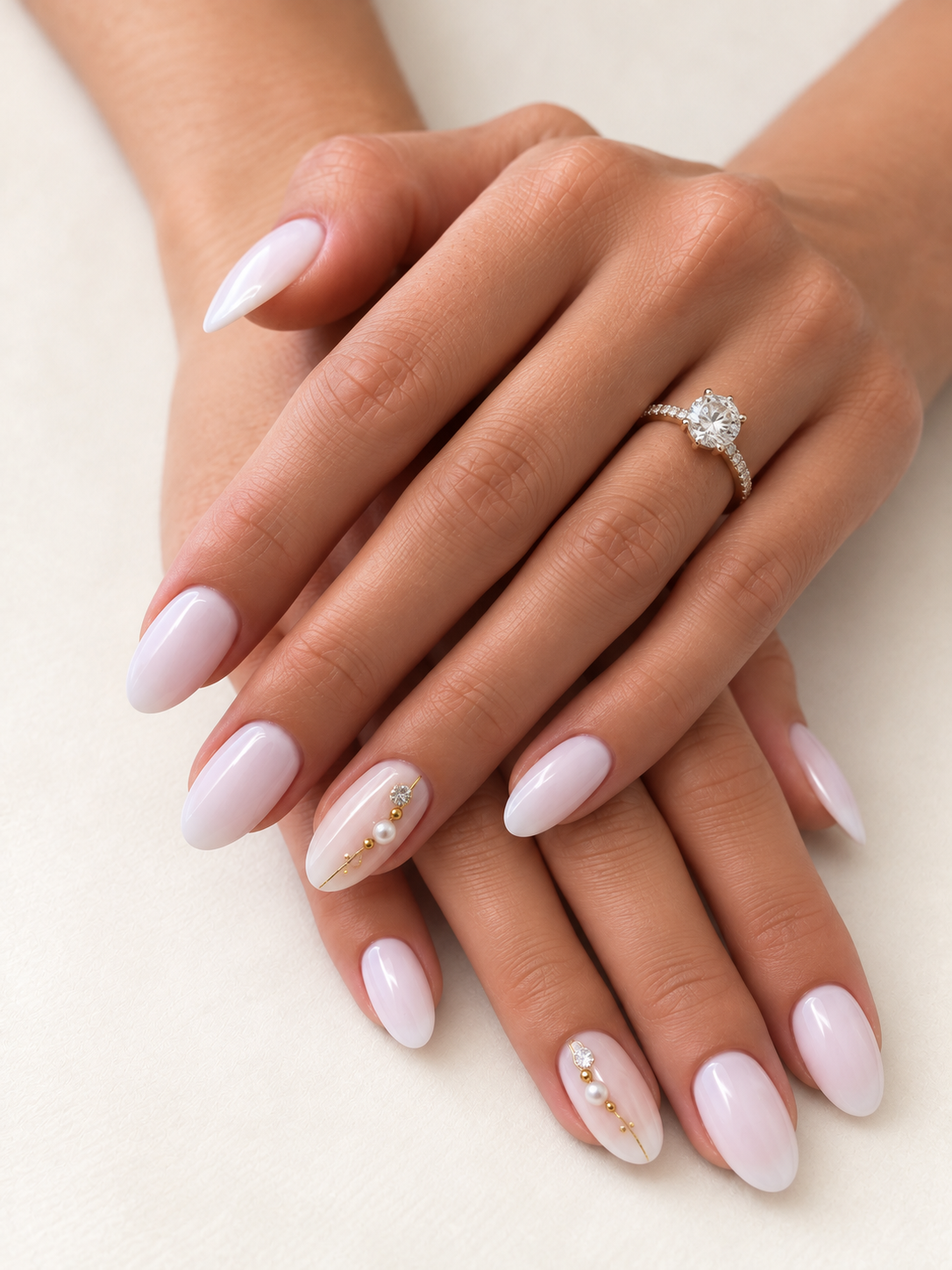 Deux mains aux ongles amande blanc nacré, ornés de fins détails dorés, perles et cristaux, création Joaillerie de l’ongle